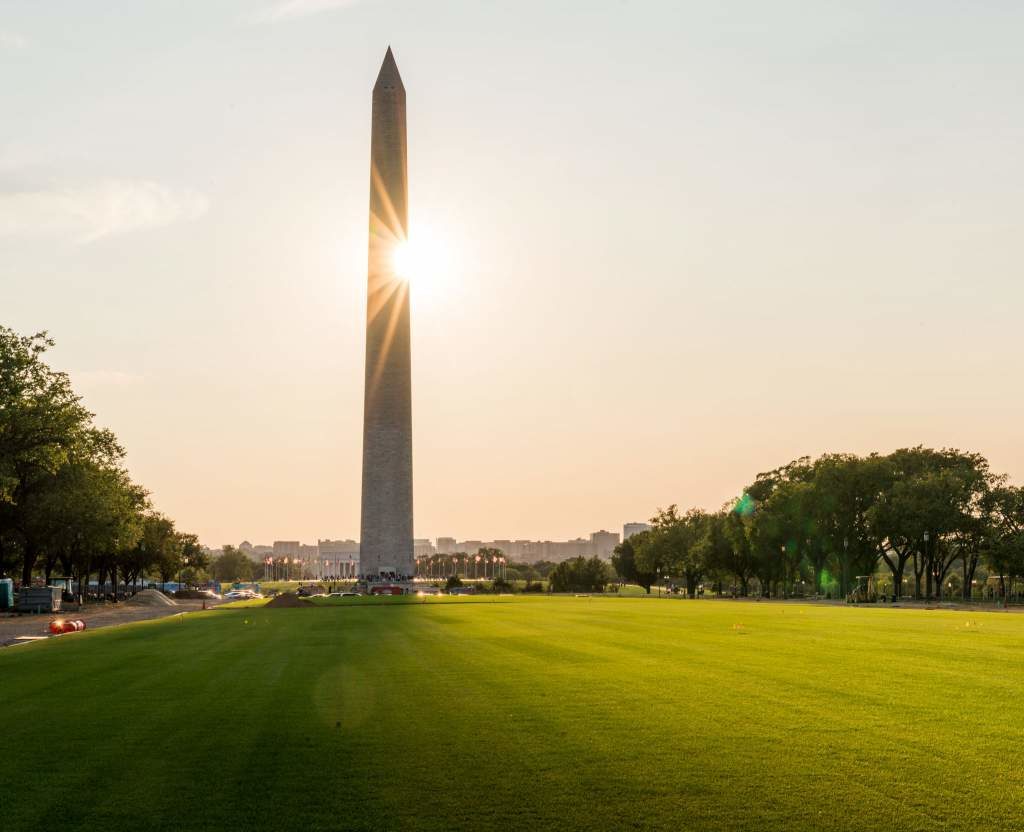 a tall monument in a grassy area with trees and a city in the background