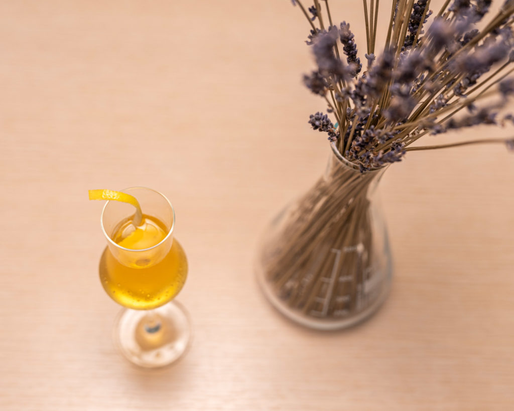 a glass of yellow liquid next to a vase of lavender
