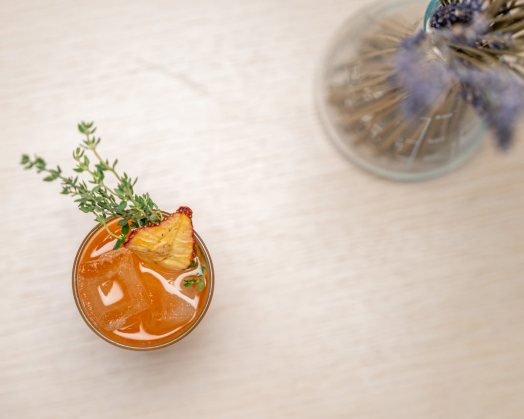 a glass of orange drink with a slice of pineapple and thyme